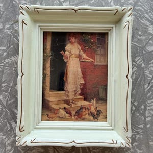 May include: A rectangular off-white frame with gold-toned accents surrounding a print of a person in a cream-colored dress standing on stone steps, holding a tray while feeding a group of chickens in front of a red brick building exterior.