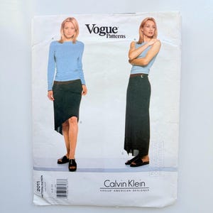 May include: A Vogue Patterns sewing pattern envelope for a Calvin Klein design, featuring a light blue long-sleeve top and a dark charcoal skirt in two lengths. The front displays two views of the garment, labeled A, B, C, and D, with the text Vogue Patterns and Calvin Klein printed on the white paper packaging.