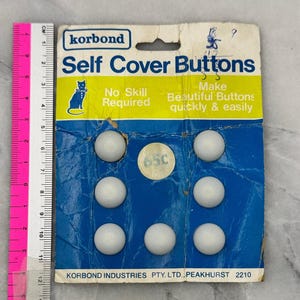 May include: A Korbond brand package of seven white self-cover buttons on a blue and yellow cardboard backing. The packaging features a cat illustration and text including "Self Cover Buttons". A pink ruler shows the package is 13.3 cm tall.