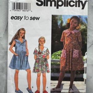 May include: Simplicity sewing pattern for girls' dresses. The pattern features illustrations of three different dress styles, including a blue dress with pockets, a floral dress, and a dress with a pink collar. The text "easy to sew" is also visible.