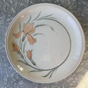 May include: A white ceramic plate with a floral design. The plate features a peach-colored iris flower and buds with green leaves and stems. The design is painted on the rim of the plate.