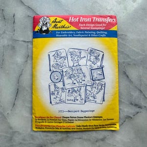 May include: Aunt Martha's Hot Iron Transfers package with a yellow background and red and blue accents. The package features illustrations of roosters and other barnyard animals. Text includes "Barnyard Happenings" and instructions for embroidery, fabric painting, and quilting.