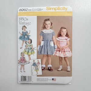 May include: Simplicity sewing pattern 8062 for children's dresses in sizes 4, 5, 6, 7, 8. The envelope features illustrations and photographs of two children wearing gingham and floral dresses with collars, pockets, and short sleeves in 1950s style.
