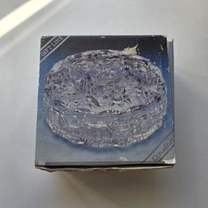 May include: A clear, round, faceted glass trinket box with a starburst pattern on the lid, presented in a dark blue cardboard box featuring the text GIFT LINE and Coretra printed in white on the diagonal corners of the packaging.