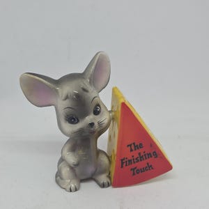 May include: A grey ceramic mouse figurine standing upright next to a triangular wedge of yellow Swiss cheese. The cheese has a red front face with the black text The Finishing Touch printed on it. The mouse has large ears and dark eyes.