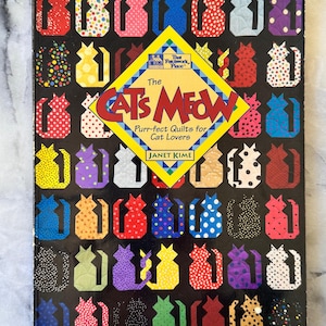 May include: A book titled "The Cat's Meow" with a black cover featuring colourful cat silhouettes. The book has a yellow diamond-shaped sticker with text. The book is about purr-fect quilts for cat lovers.