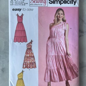 May include: Simplicity sewing pattern S9746 with three dress designs. The pattern includes a pink one-shoulder dress, a tiered floral dress, and a yellow dress. The pattern is easy-to-sew and designed and printed in the USA.
