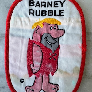 May include: Embroidered patch featuring Barney Rubble from The Flintstones cartoon. The patch has a white background with a red border. Barney is depicted in pink and red with yellow hair and the text "BARNEY RUBBLE" in black.