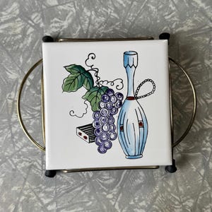 May include: Square ceramic trivet with a hand-painted design featuring a bunch of purple grapes, green leaves, and a blue bottle. The trivet is set in a gold-toned metal frame with black feet.