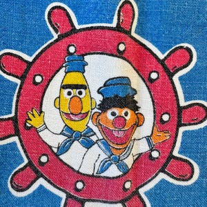 May include: A blue fabric featuring a graphic print of Bert and Ernie dressed in white sailor outfits with blue neckerchiefs. They are positioned inside a red ship's wheel with white circular accents, set against a solid blue background.