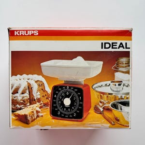 May include: A vintage red and white KRUPS kitchen scale with the word "IDEAL" on the box. The scale has a white bowl filled with flour and a black dial. The box also shows a cake and other baking tools.