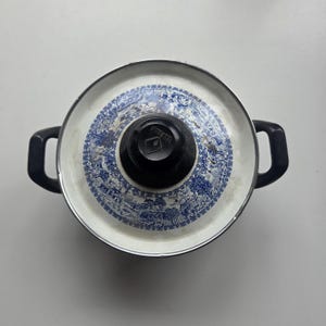 May include: A round metal cooking pot with a lid featuring a blue and white transferware pattern of landscapes and trees. The pot has two black side handles and a central black knob on the lid with the embossed text TOUTEN visible on the top.