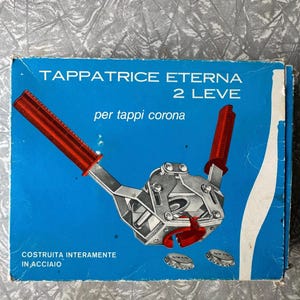 May include: A vintage bottle capper in a blue box. The capper is silver with red handles. The box has the text "Tappatrice Eterna 2 Leve" and "per tappi corona". The box also says "Costruita interamente in acciaio".