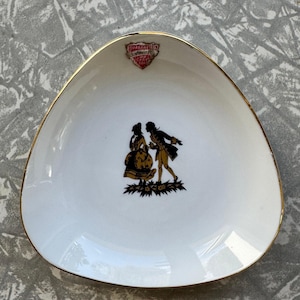 May include: White, triangular-shaped dish with a gold rim. The dish features a black and gold silhouette of a couple and the text "GUARANTEED" in a red and gold crest. A decorative piece.