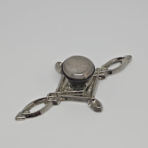 Puede incluir: Un pomo de metal plateado para muebles con una parte superior circular y una placa trasera en forma de diamante con extremos puntiagudos y calados. El herraje tiene un acabado pulido con un poste central que une el pomo a la base.