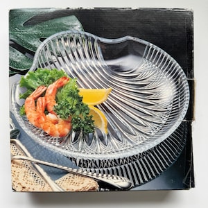 Könnte beinhalten: Eine Servierplatte aus klarem Glas mit einem einzigartigen, gewellten Design. Die Platte ist mit gekochten Garnelen, frischer Petersilie und Zitronenscheiben gefüllt. Die Platte befindet sich auf einer Schachtel mit schwarzem Hintergrund und einem Blattmuster.