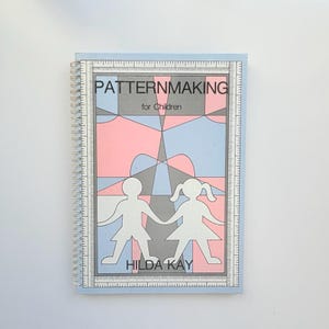 Op de afbeelding: Een spiraalgebonden instructieboek getiteld Patternmaking for Children door Hilda Kay. De kaft heeft een geometrisch ontwerp in roze, blauwe en grijze tinten, met witte silhouetten van twee kinderen en een rand die op een liniaal lijkt.