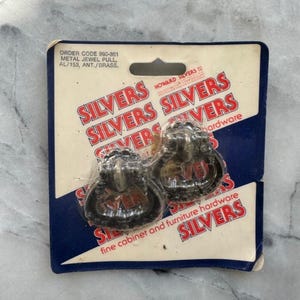 May include: Two antique-style cabinet pulls on a cardboard backing. The pulls are dark metal with a decorative design. The packaging reads "SILVERS" in red and white, and "fine cabinet and furniture hardware."
