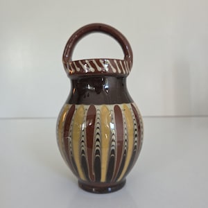 May include: A small ceramic vessel with a single arched handle at the top. The body features a repeating vertical pattern of brown, tan, and black marbled stripes. A decorative band with diagonal white lines encircles the neck of the vessel.