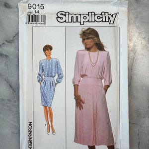 May include: A vintage Simplicity sewing pattern packet. The packet features illustrations of two dresses, one blue with white polka dots and one pink. The packet has the number 9015 and the word "Simplicity" printed on it.