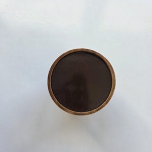 Puede incluir: Un objeto circular que presenta una superficie central plana, lisa y de color marrón oscuro, rodeada por un borde fino de madera de color marrón claro. El objeto se ve directamente desde arriba, mostrando su diseño simple, redondo y simétrico con un contraste claro entre el interior oscuro y el borde de madera más claro.