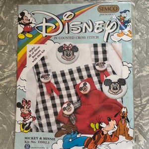 May include: A Disney counted cross stitch kit package featuring Mickey and Minnie Mouse, Donald Duck, Goofy, and Pluto. The packaging displays finished cross-stitched buttons with character faces, a black and white gingham garment, and red fabric.