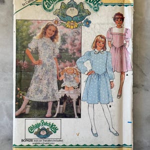 May include: Vintage Butterick sewing pattern for Cabbage Patch Kids outfits. The pattern features illustrations of girls in dresses, with the Cabbage Patch Kids logo and text. Includes a free pattern for one outfit and iron-on transfers.