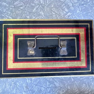 Può includere: Scatola rettangolare in metallo nero e oro con manico e fessura. La scatola ha una striscia rossa e un bordo dorato. Il manico e la fessura sono centrati sulla parte superiore della scatola. La scatola è su una superficie azzurra.