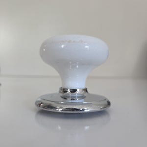 May include: A white ceramic cabinet knob with a smooth, rounded top and a tapered neck. The base of the knob features a polished, reflective chrome-colored metal circular plate that connects to the white ceramic body, creating a clean contrast.