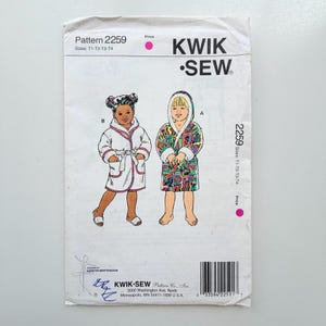 May include: A Kwik Sew pattern envelope labeled 2259 for sizes T1-T4. The front shows illustrations of two children wearing hooded robes: one in a solid white robe with pink trim and one in a multi-colored patterned robe with white trim.