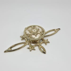 Könnte beinhalten: Ein goldfarbener Schubladengriff aus Metall mit einem runden Knopf, der ein zentrales Blumenmotiv und eine weiße Einlage aufweist. Der Knopf ist auf einer länglichen, dekorativen Rückplatte mit symmetrischen, durchbrochenen Schnörkeln montiert.