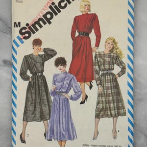 Pode incluir: Padrão de costura Simplicity vintage, tamanho 12 Miss, apresentando quatro modelos de vestidos. A capa tem um fundo branco com bordas listradas em azul e branco e o logotipo Simplicity. Inclui uma tabela especial sobre xadrez.