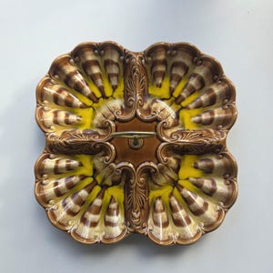 May include: A square ceramic serving dish with four partitioned sections, featuring a glossy brown and yellow glaze. The dish has a scalloped edge and a central metal handle. The surface is decorated with embossed, shell-like radial patterns.