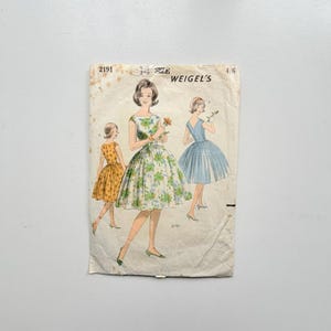 May include: A vintage Weigel's sewing pattern envelope, number 2191, size 14. The paper envelope features illustrations of three dresses: one yellow with a floral print, one white with a green floral pattern, and one solid light blue dress.