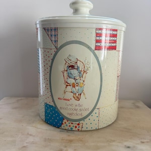Pode incluir: Um frasco de biscoitos vintage Holly Hobbie com tampa e corpo creme. O frasco apresenta um design de patchwork com detalhes em vermelho, azul e branco. Um gráfico oval mostra Holly Hobbie com o texto "Love is the good cooks secret ingredient."