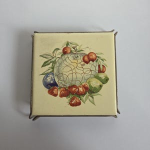 May include: A square ceramic tile trivet featuring a central illustration of a large melon surrounded by strawberries, plums, and green leaves on a cream background, held within a simple, thin, dark metal wire frame stand.