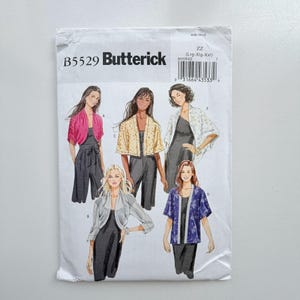 May include: A Butterick B5529 sewing pattern envelope featuring illustrations of five different styles of open-front jackets or shrugs worn over black jumpsuits. The designs include varying sleeve lengths, patterns, and solid colors like pink, white, and blue.