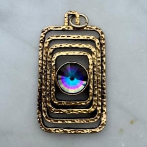 May include: A gold-toned pendant with a rectangular, layered design. The pendant features a central, iridescent, round gemstone that reflects multiple colors. The metal has a textured, hammered appearance, and a small loop is at the top for a chain.