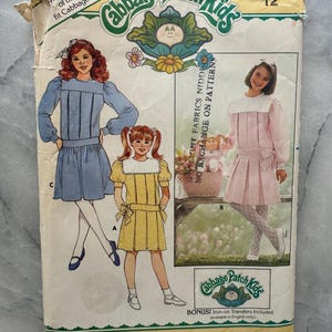 May include: Vintage Butterick sewing pattern for Cabbage Patch Kids outfits. The pattern features illustrations of children's dresses in blue, yellow, and pink. The pattern size is 12. The packaging is yellowed with age and includes the text "Cabbage Patch Kids".