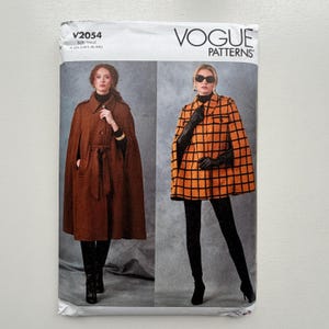 May include: Vogue Patterns sewing pattern featuring two women modeling capes. One cape is brown with a belt, the other is orange and black checkered. The pattern number is V2054.