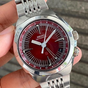 May include: A vintage Seiko automatic wristwatch with a stainless steel strap and case. The watch has a deep red dial with white hands and markers. The dial includes the text "SEIKO AUTOMATIC".