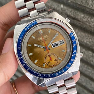 May include: Vintage Seiko Chronograph automatic watch with a silver stainless steel band. The watch face is gold with a blue and red bezel. The watch displays the day and date.