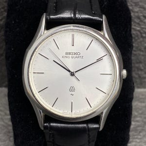 Può includere: Un orologio Seiko King Quartz argentato con quadrante bianco e cinturino in pelle nera. Il quadrante dell'orologio presenta lancette e indicatori neri, con il nome del marchio e il logo. Design classico e minimalista.