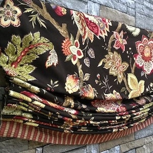 May include: A decorative Roman shade featuring a dark brown floral pattern with red, yellow, and green accents. The fabric is folded in a layered design, with a striped border at the bottom. The shade is mounted on a stone wall.