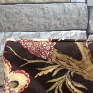 May include: Close-up of a brown fabric with a floral pattern in red, yellow, and cream. The fabric has a dark brown background with a tree branch design. The fabric is likely used for upholstery or home decor.