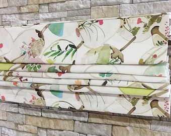 May include: White roman shade with a watercolor print of green leaves, pink and blue flowers, and brown branches. The shade is folded up, showing the layers of the fabric.