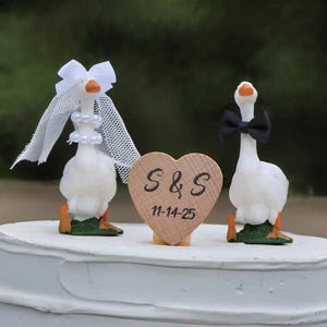 May include: Wedding cake topper featuring two white goose figurines. One goose wears a veil and pearl necklace, the other a black bow tie. A wooden heart with "S&S" and the date 11-14-25 is between them. The cake is white.