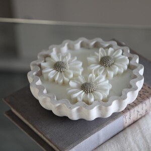 May include: A white daisy-shaped candle with three flower accents, set in a scalloped dish. The candle sits atop a stack of books, creating a serene, decorative display. The candle is a cream color.