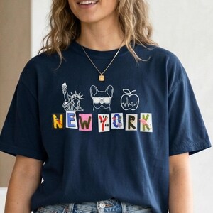 May include: Navy blue t-shirt with a New York City theme. The shirt features white line drawings of the Statue of Liberty, a dog wearing sunglasses, and an apple with a heartbeat. The word "NEW YORK" is spelled out in colorful, block letters.
