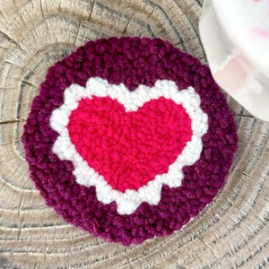 May include: A handmade, round coaster with a heart design. The coaster features a bright pink heart in the center, surrounded by a white scalloped border and a deep purple outer edge. The coaster is made of a textured, crocheted material.